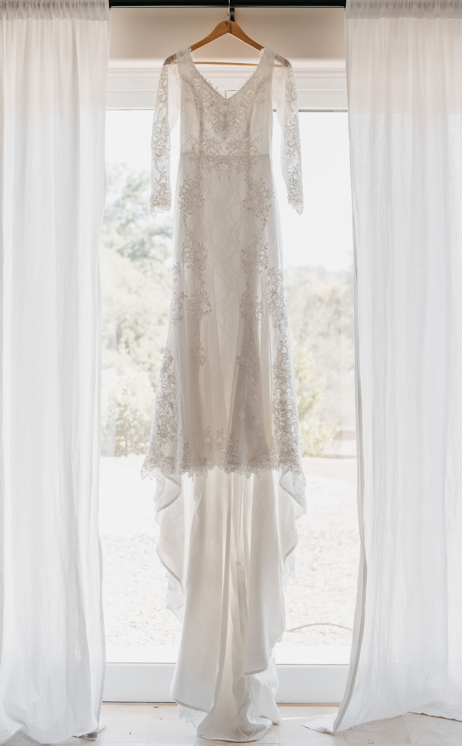 An elegant white lace wedding dress hangs in front of a bright window with flowing curtains.