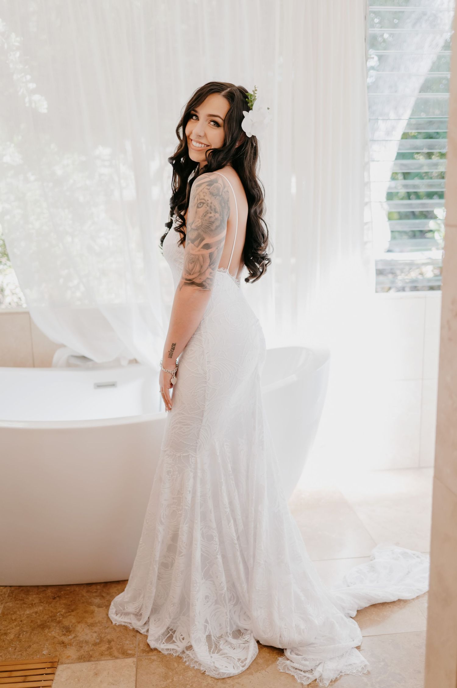 A bride in a fitted lace gown poses elegantly against white curtains in natural light.