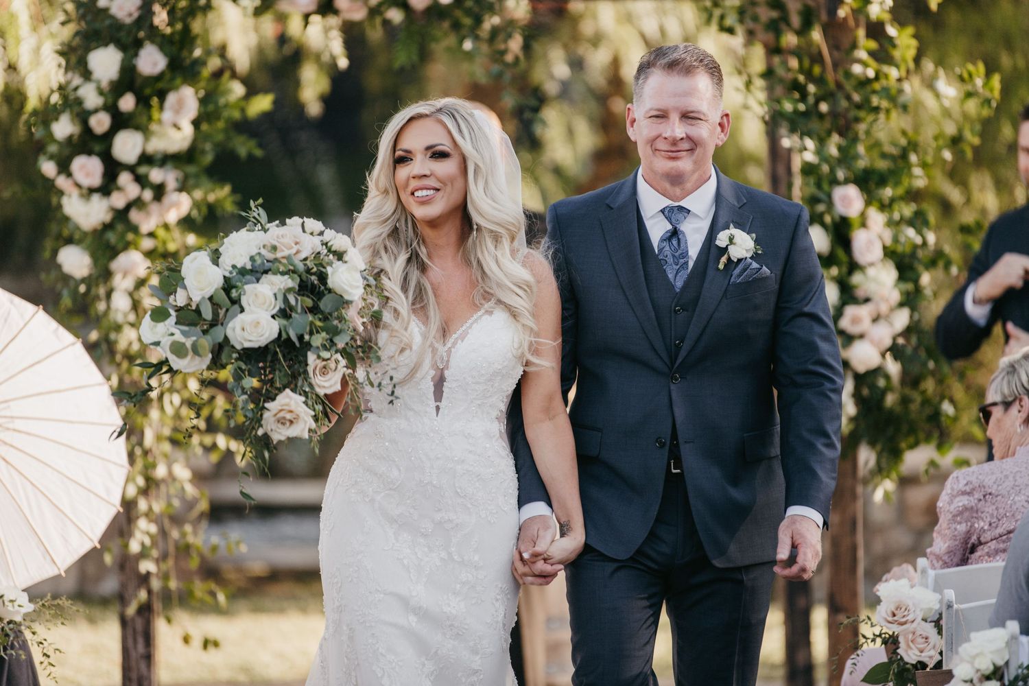 A wedding couple walks hand in hand under floral decorations during their outdoor ceremony.