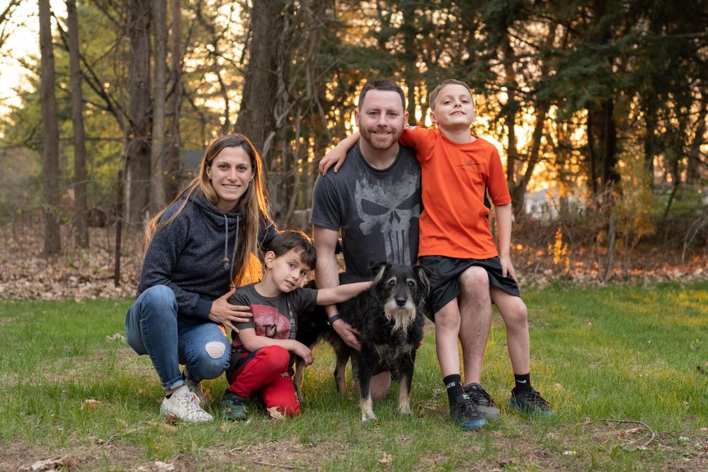 Family with a black and white dog poses together on grass during golden hour sunset in wooded setting.