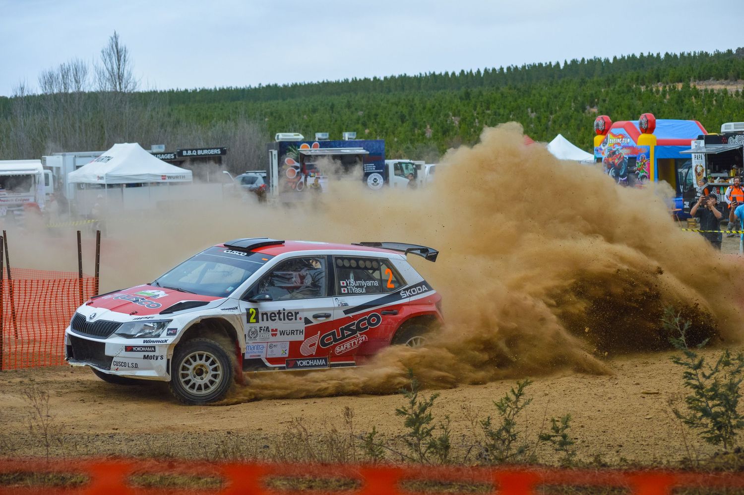 Rally car kicks up dramatic dust cloud while racing on dirt track.