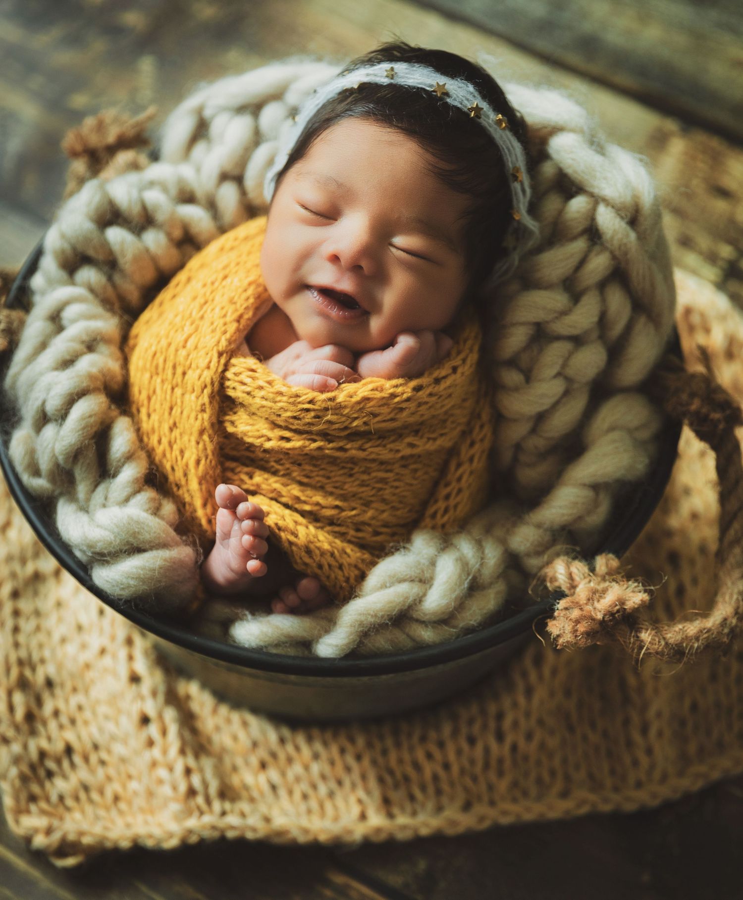 Smiling newborn wrapped in mustard yellow knit blanket nestled in a metal bowl with chunky yarn.