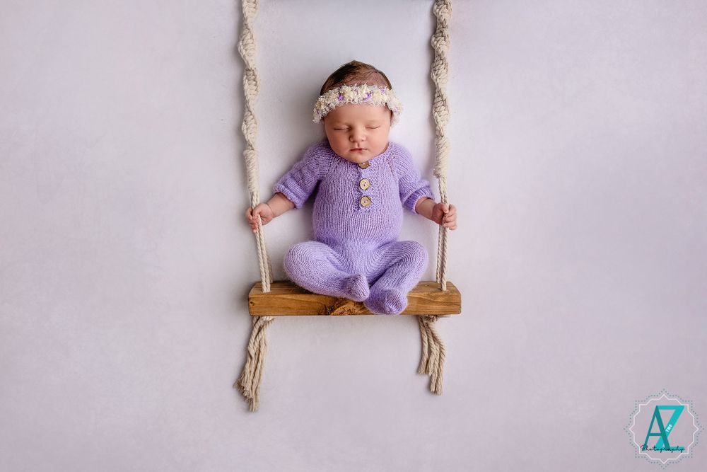 Baby wearing purple romper sitting on wooden rope swing against light background.