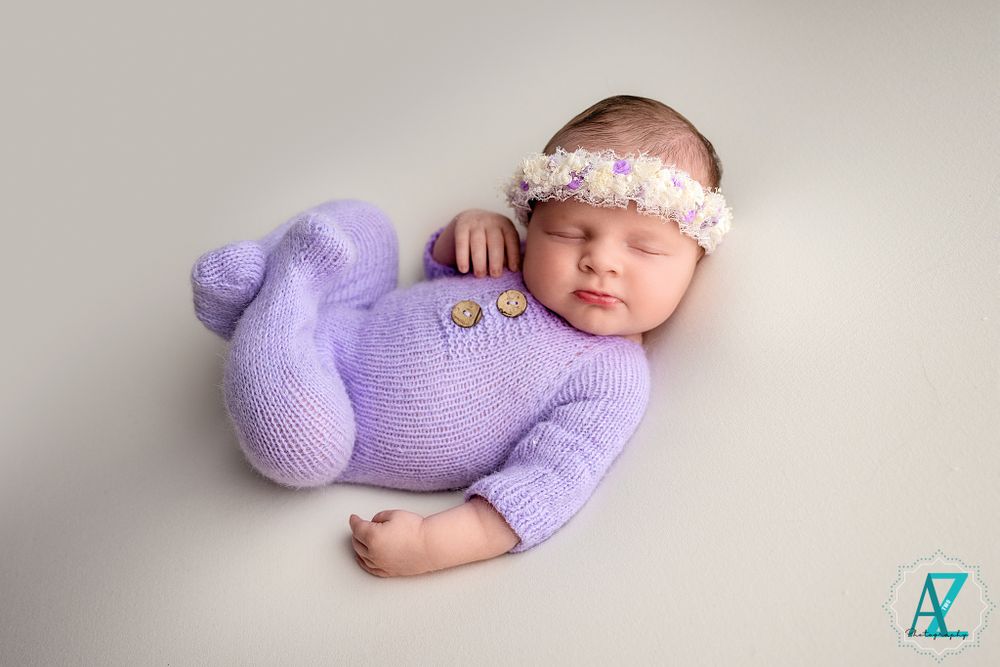 Sleeping newborn in purple knit outfit with lace headband on neutral background.