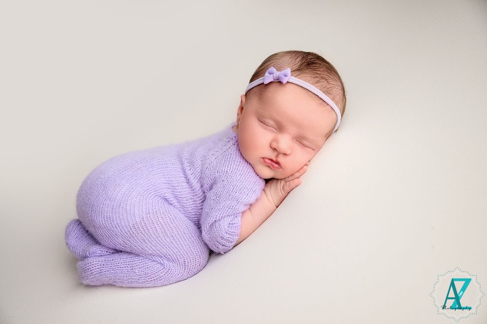 A sleeping newborn in a lavender knit outfit with a delicate headband rests peacefully on a light background.