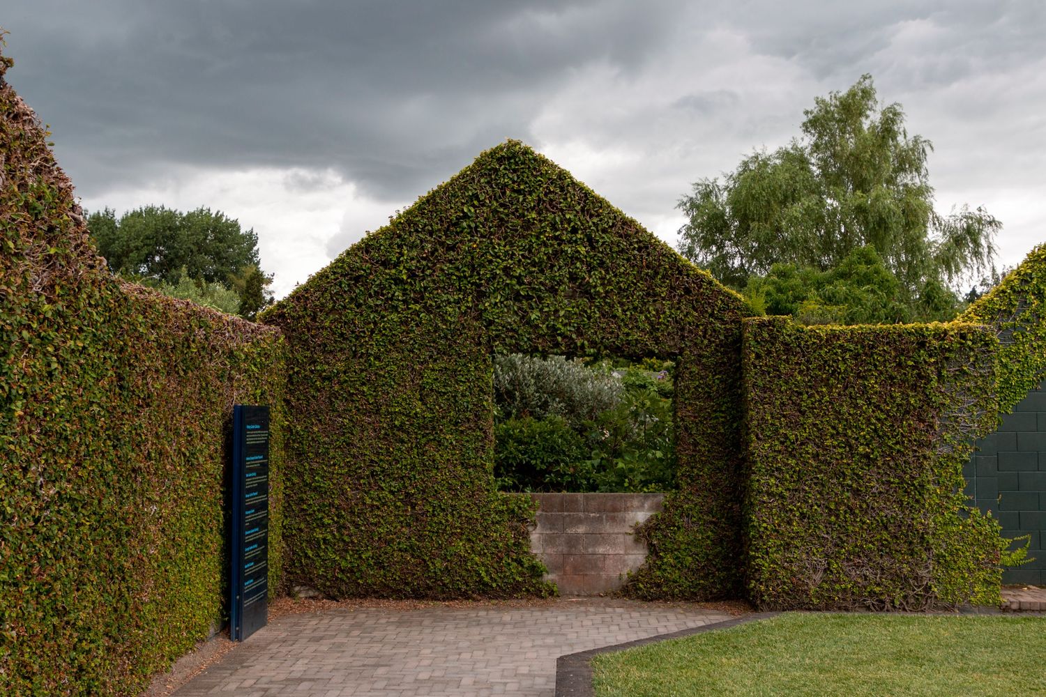 Neatly trimmed hedges form a series of green archways leading to a small open courtyard, with a dark blue gate nestled in one corner.