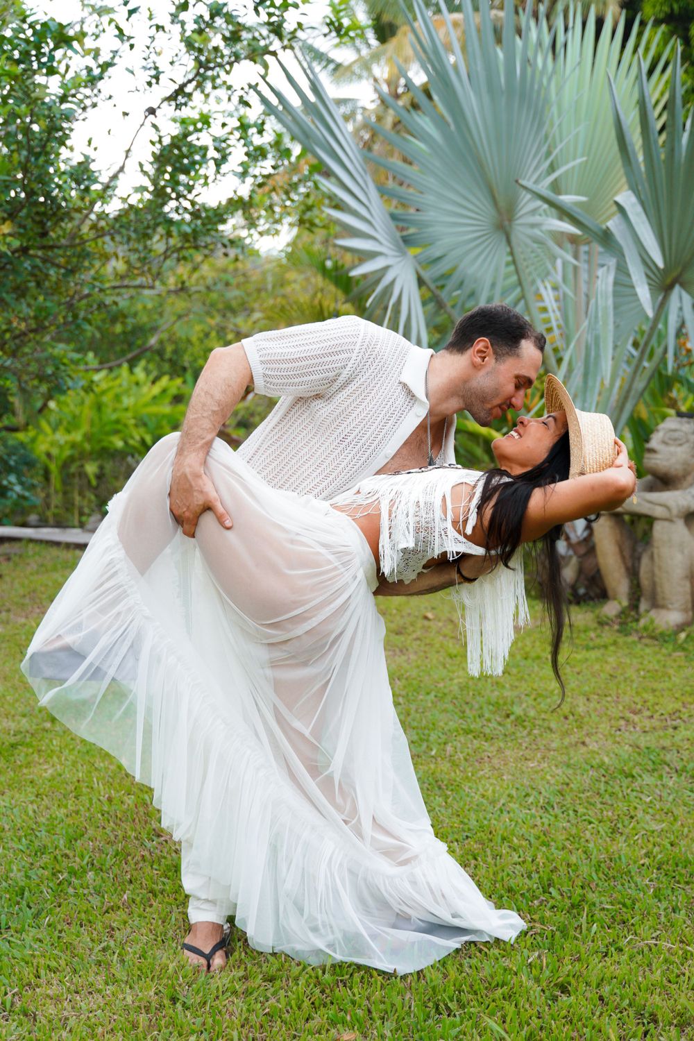 Romantic dip pose of couple in white clothing against tropical palm background.