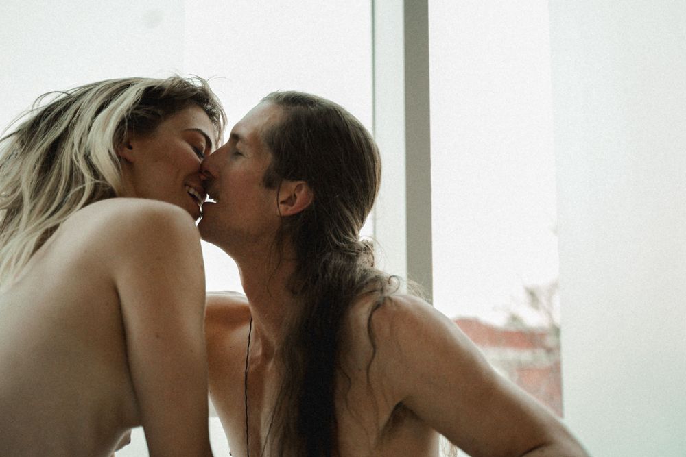 Two intimate partners share a tender kiss by natural window light in a romantic moment.