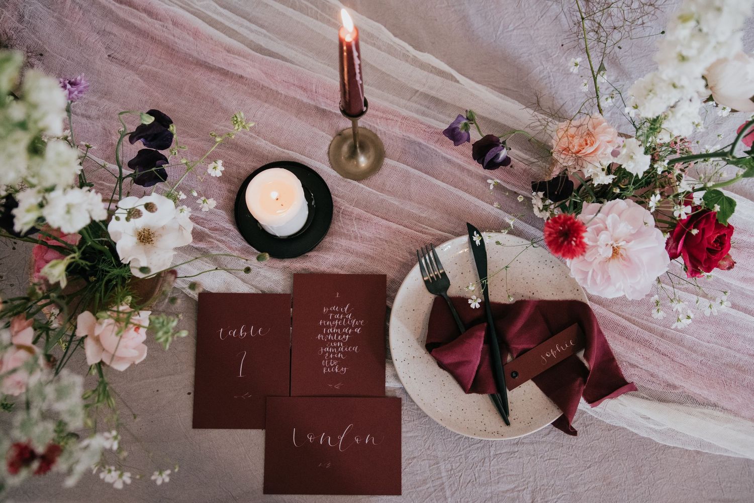 Wedding details flatlay with burgundy stationery, candles, and scattered flowers on pink fabric.