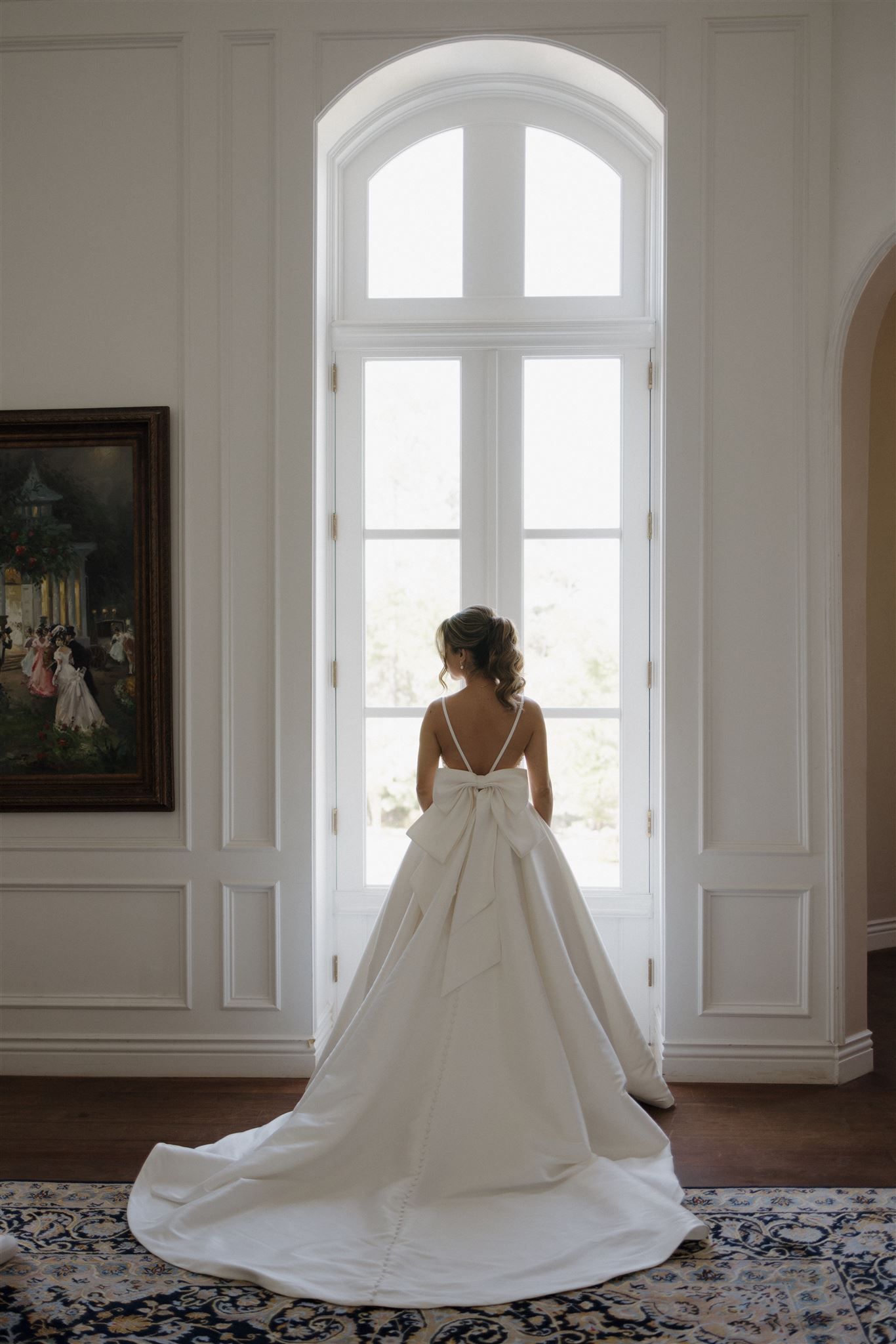 A bride in a stunning white ballgown with long train stands silhouetted against a tall arched window in an elegant room.