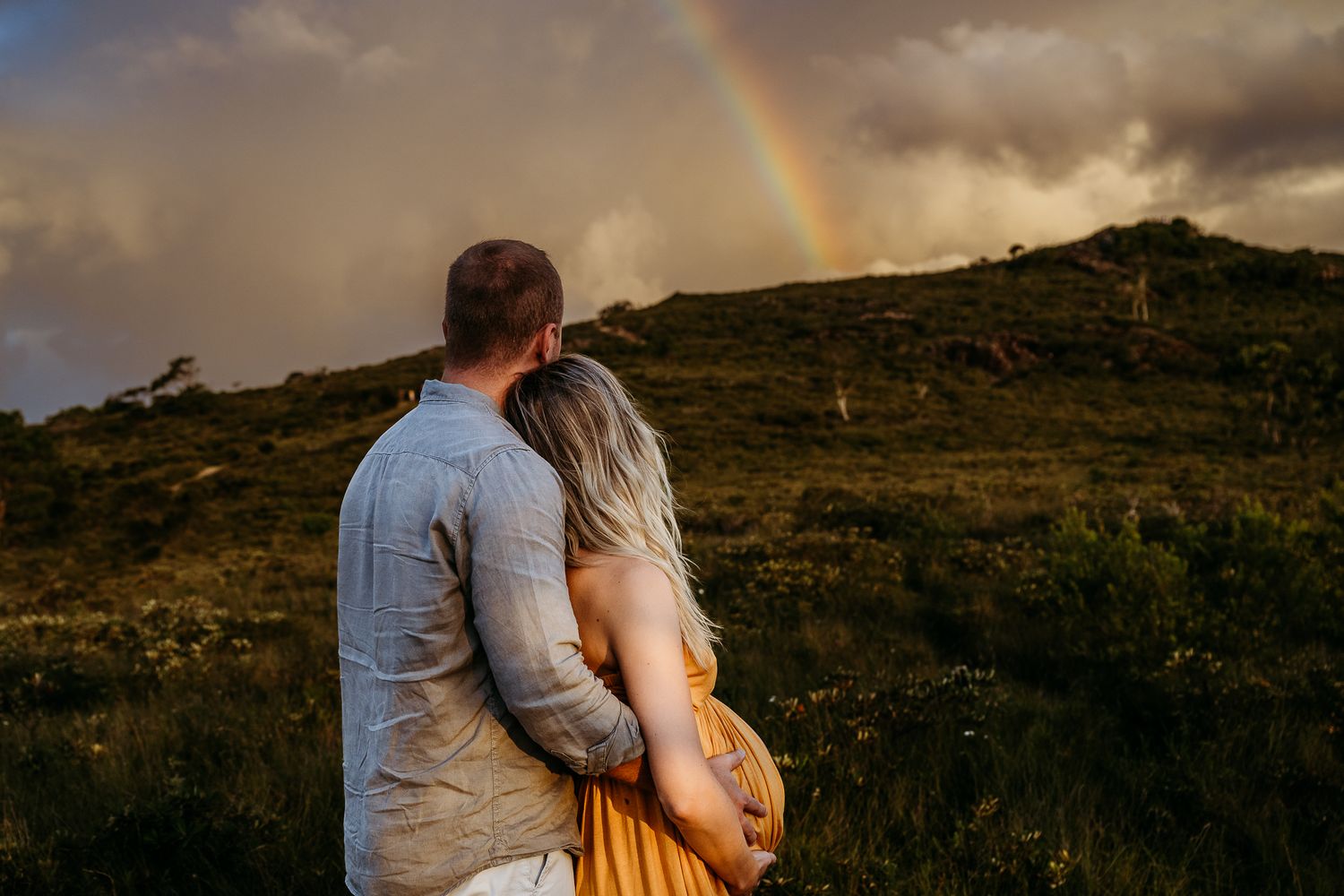A couple admires a rainbow over hills during sunset while embracing.