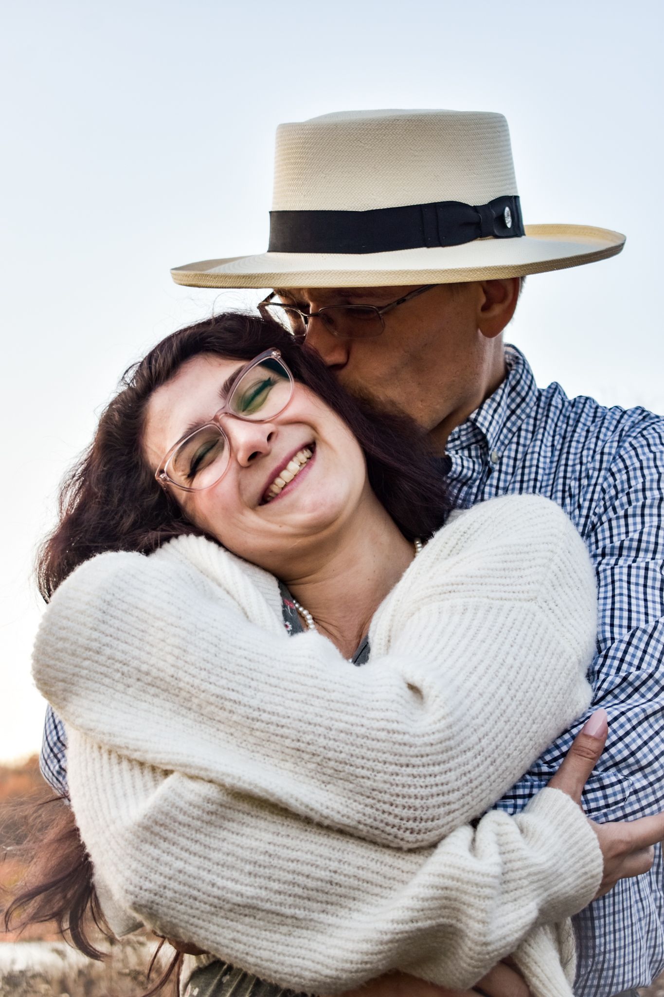 Loving couple embracing and laughing outdoors, with one wearing a white sweater and sun hat.