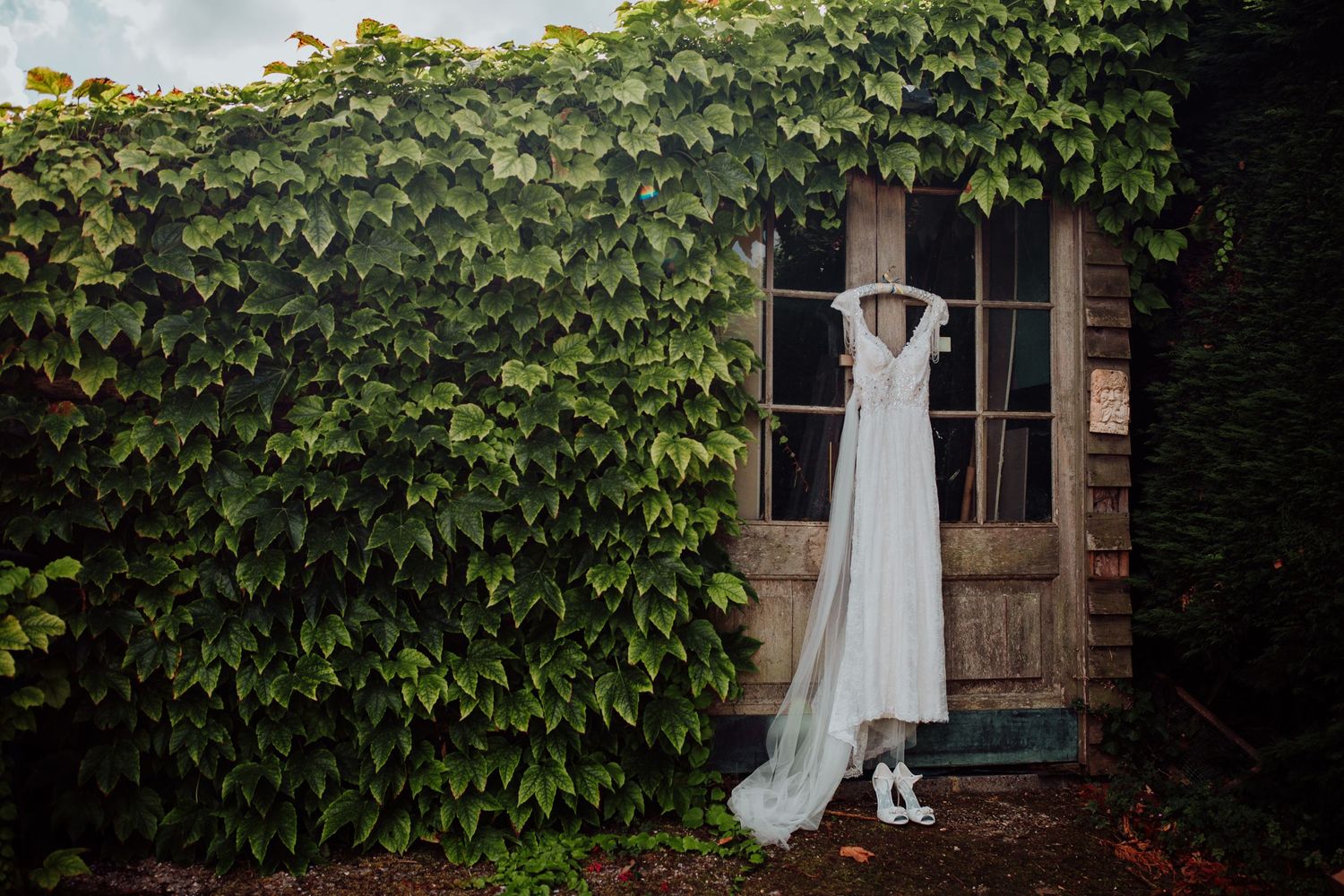 White wedding dress hanging on rustic wooden door surrounded by ivy covered walls.