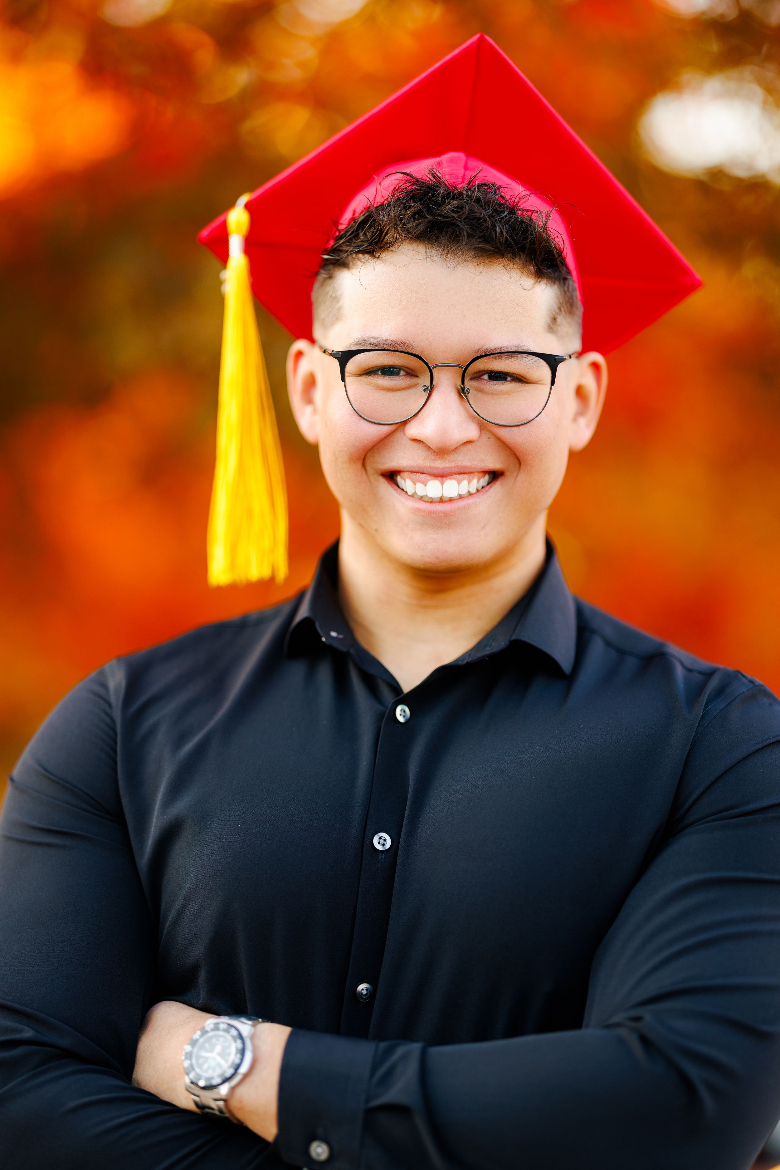 Graduate in red cap and black polo shirt smiling confidently with fall colors in background.