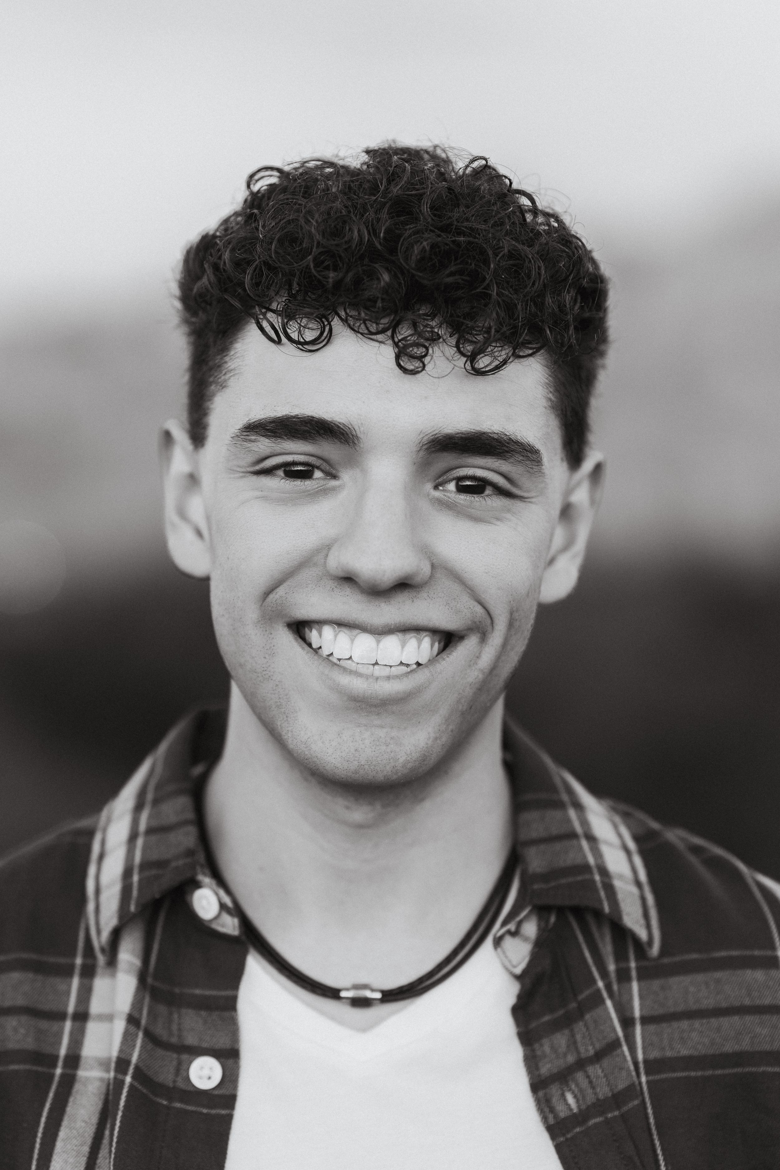 Black and white portrait of someone with curly dark hair and plaid shirt smiling brightly.