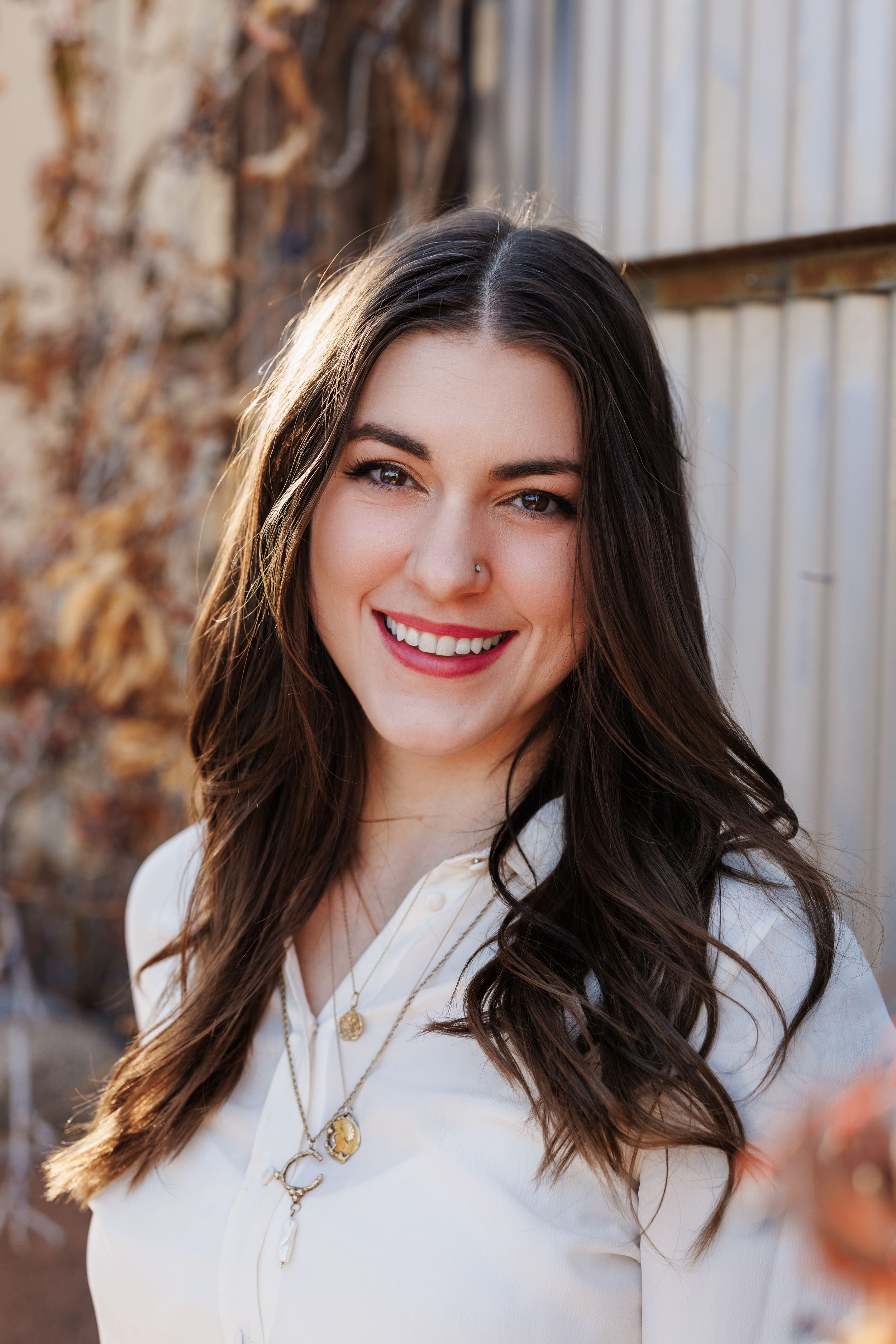 Professional headshot of someone smiling warmly in a white top with long dark wavy hair and delicate necklaces.