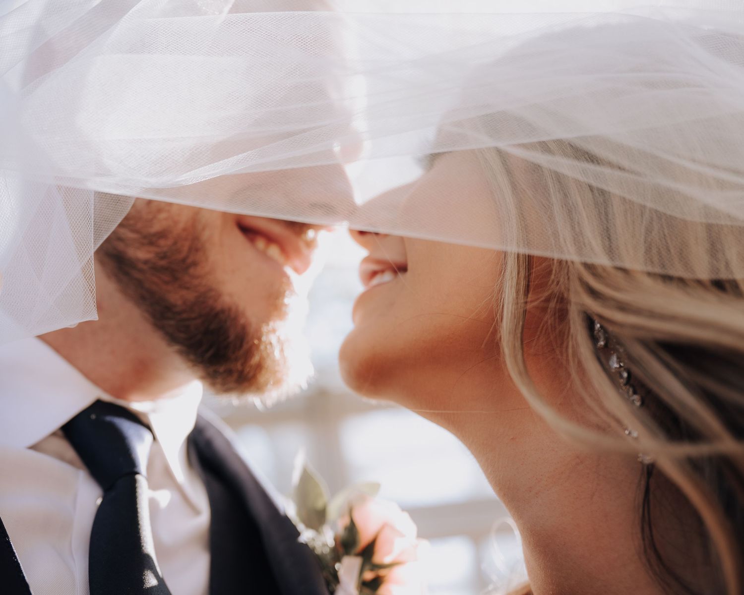 Intimate moment between newlyweds under a white veil with soft natural lighting.