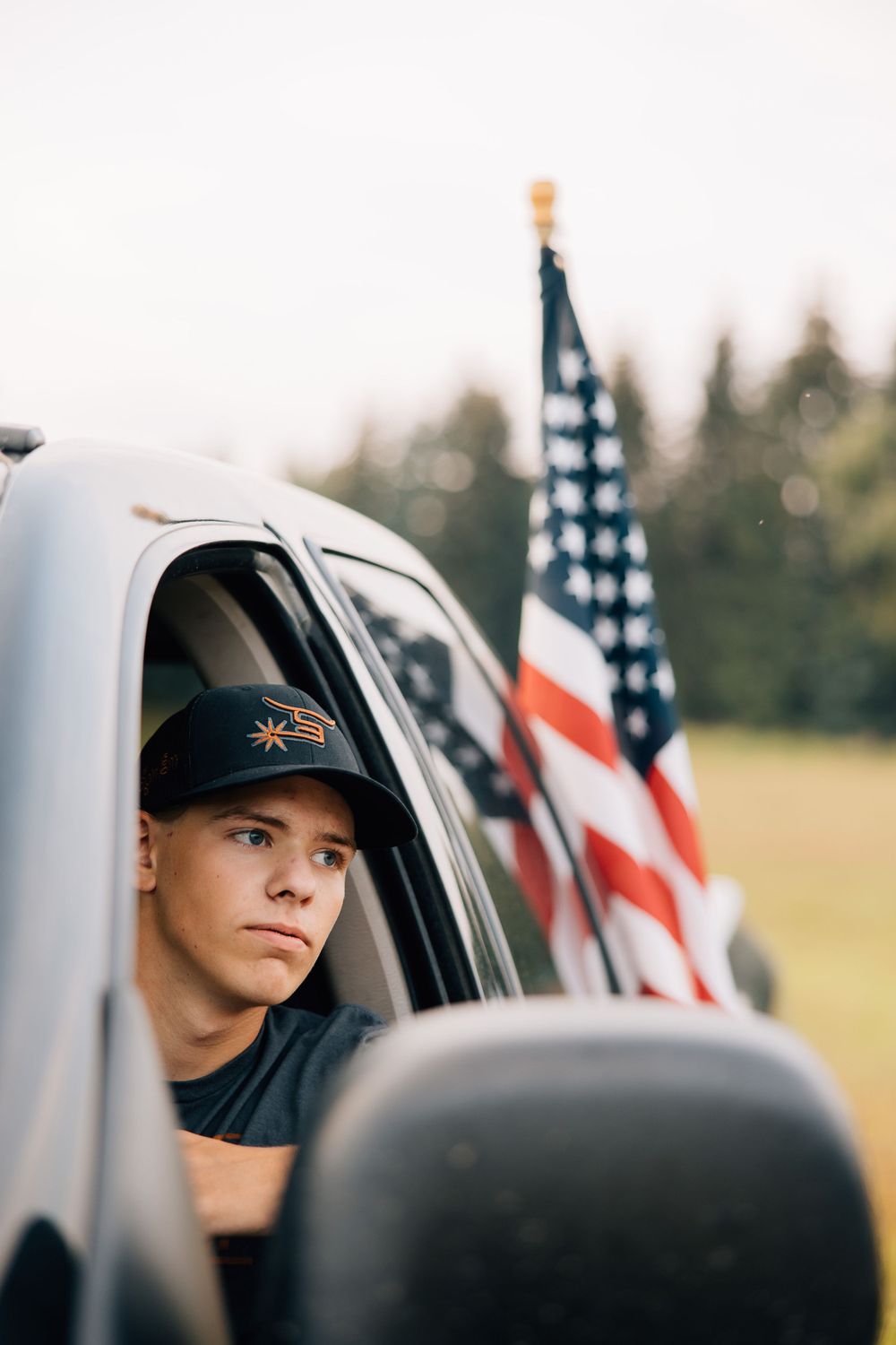 Person in baseball cap looks out truck window with American flag mounted behind.