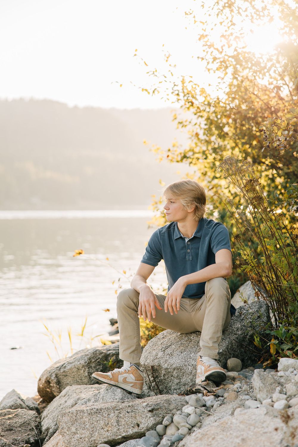Person seated on rocky shore during golden hour beside tranquil lake.