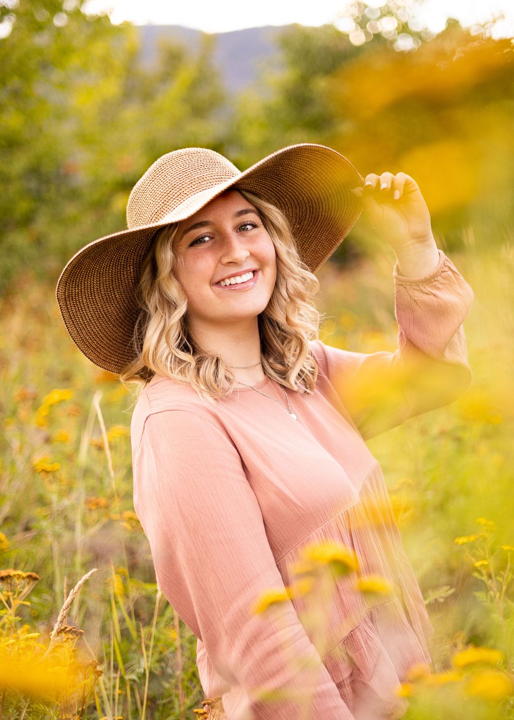 Person in wide-brim sunhat and pink sweater smiling in sunlit wildflower field.