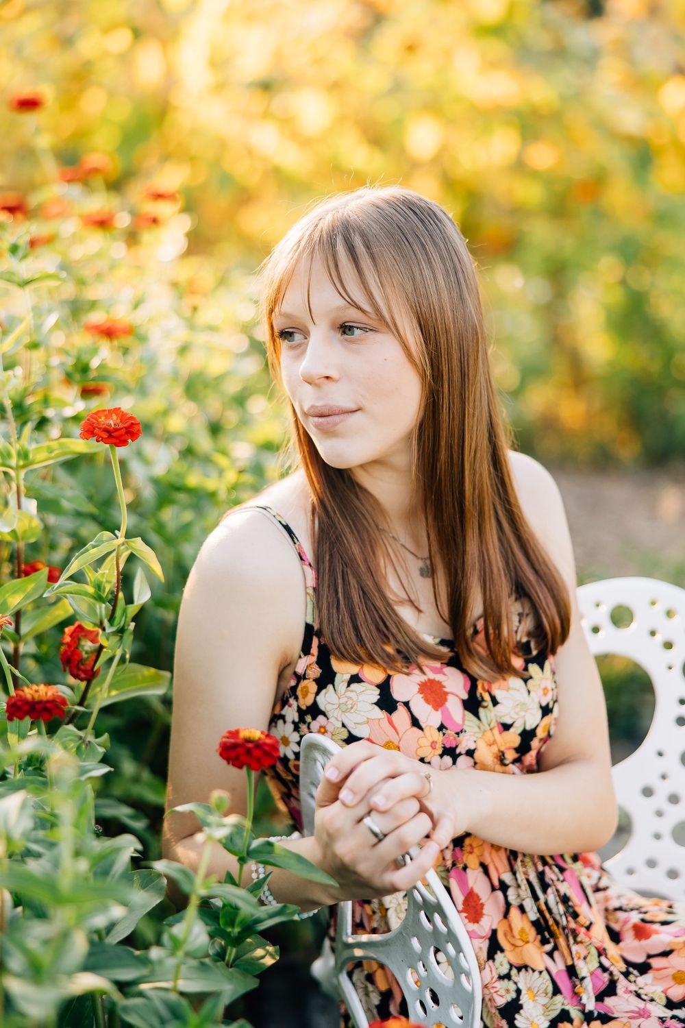 Woman in floral dress sits among red zinnias in a garden on a sunny afternoon.
