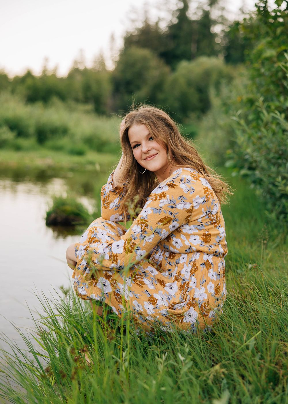 Woman in floral dress sits by a serene pond surrounded by lush greenery.