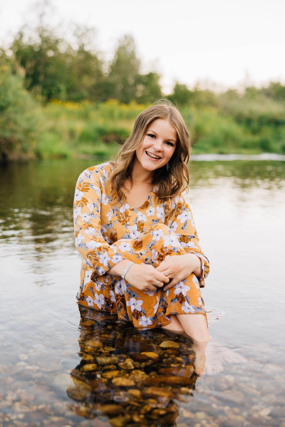 Woman in yellow floral dress sits with feet in clear water, smiling at sunset.