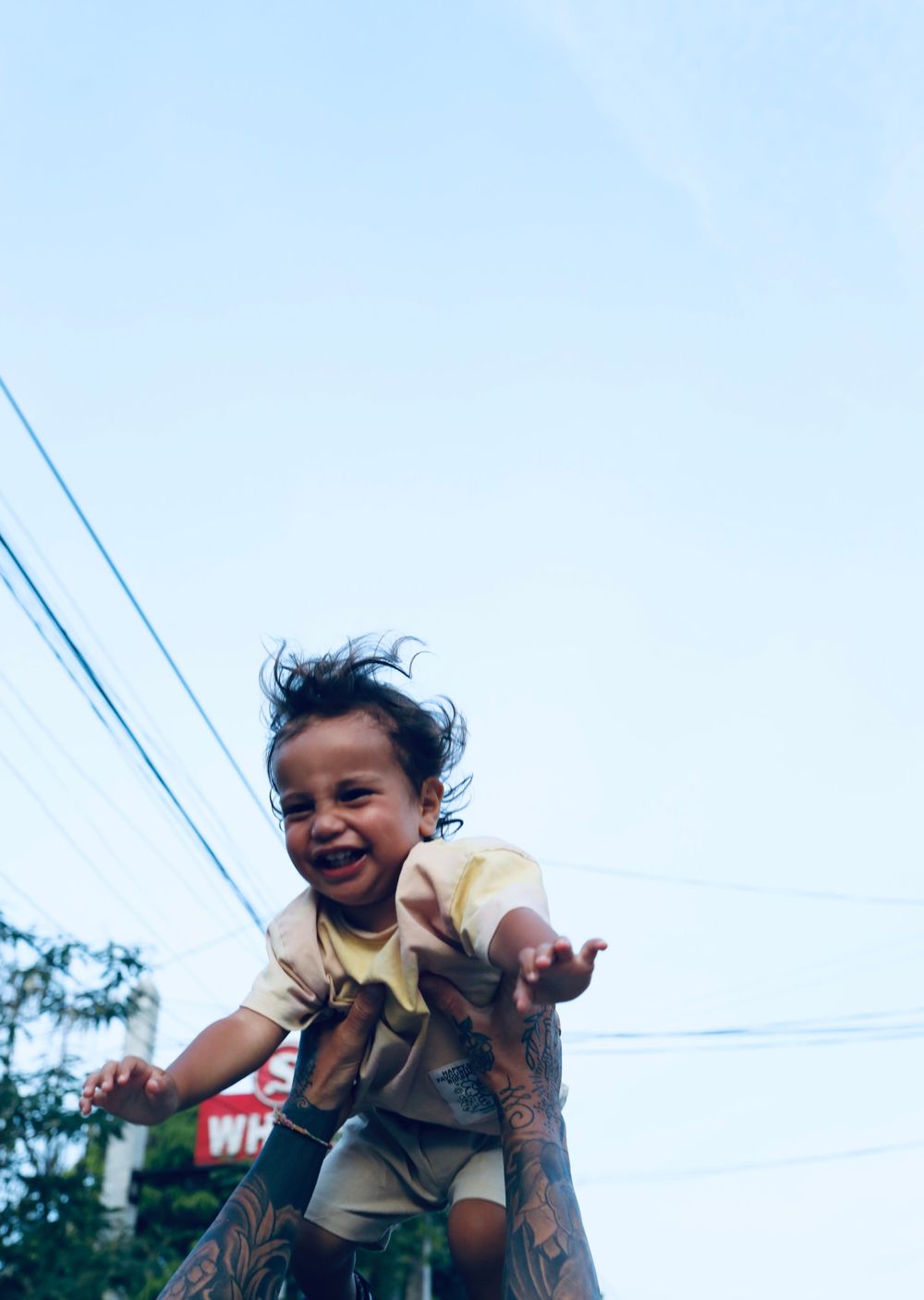 Child being lifted joyfully into bright blue sky with power lines visible in background.
