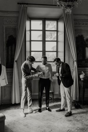 Three people gather by a tall window in formal attire during wedding preparations.