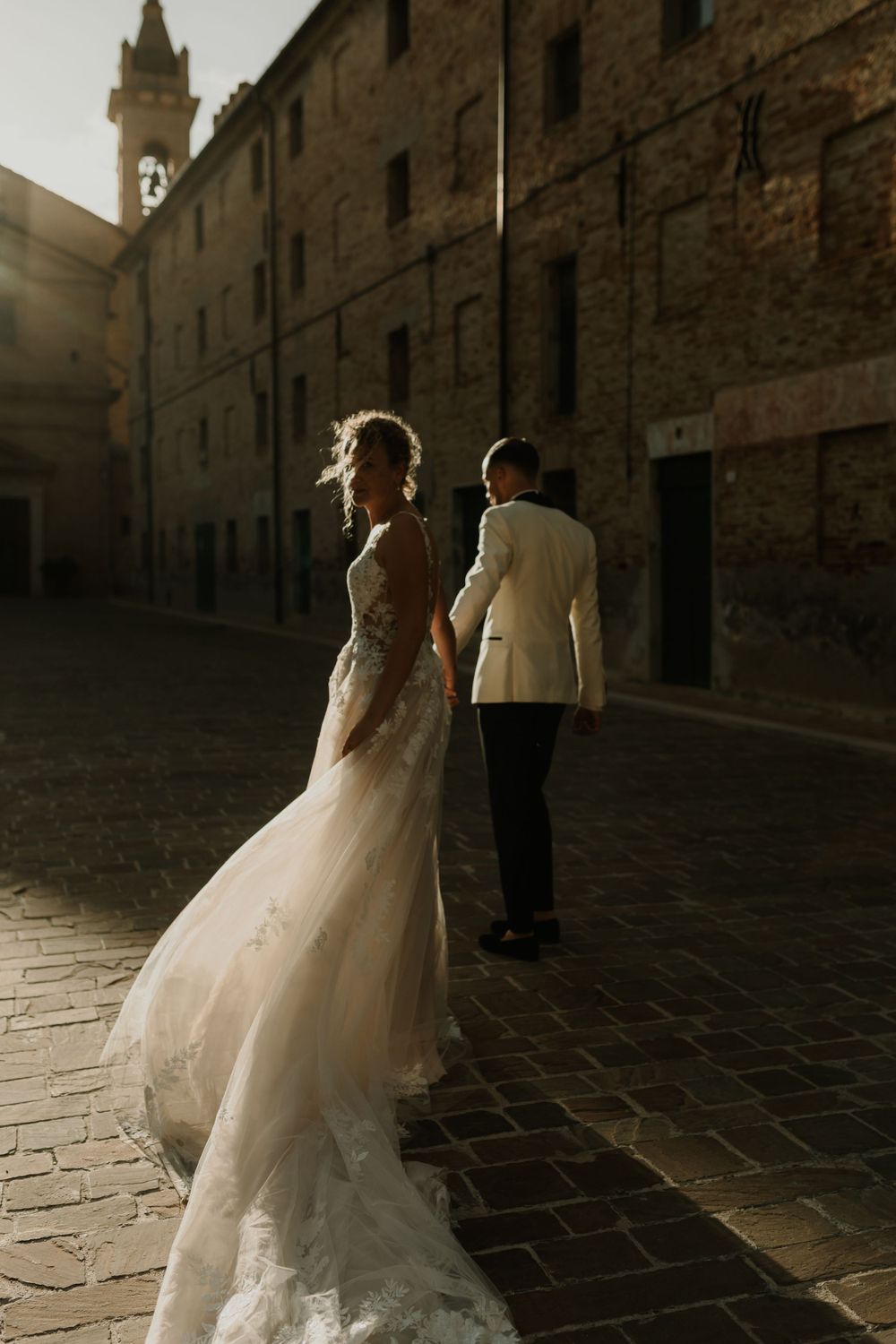 A bride and groom walk along a historic stone building in dramatic sunset lighting.