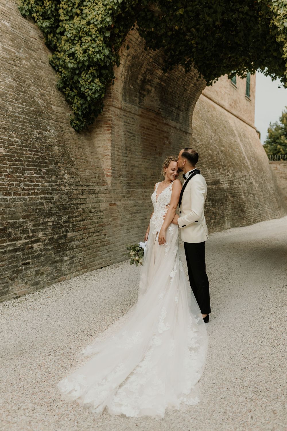 A wedding couple poses against an ivy-covered stone wall in elegant attire.