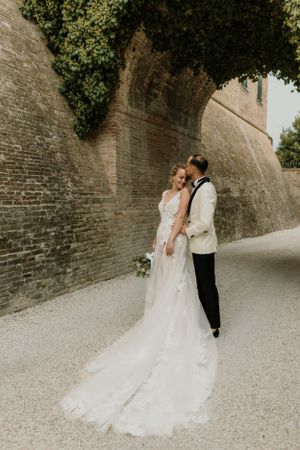 A wedding couple poses against an ivy-covered stone wall in elegant attire.