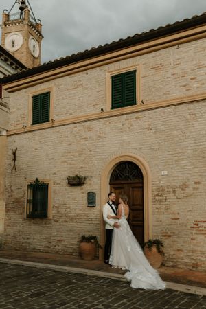 A wedding couple embraces outside a rustic Italian building with a clock tower.