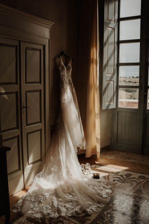 A delicate lace wedding dress hangs by a window in golden sunlight.
