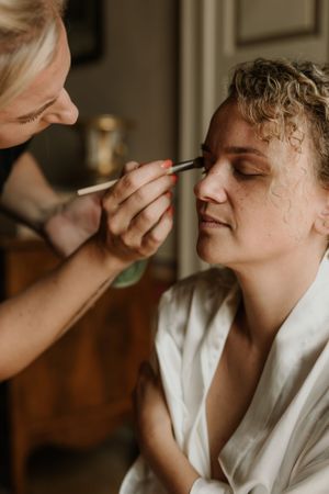 A makeup artist applies cosmetics during wedding preparation in warm natural light.