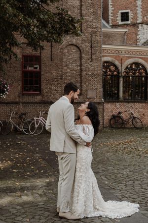 Romantic wedding couple embracing on cobblestone street with historic brick building backdrop.