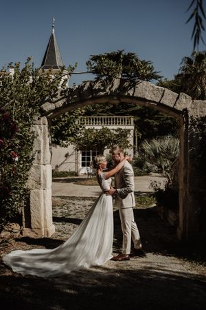 Wedding couple sharing intimate moment under garden archway with church spire in background.