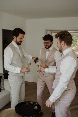 Groomsmen in light colored suits sharing drinks during wedding preparations.