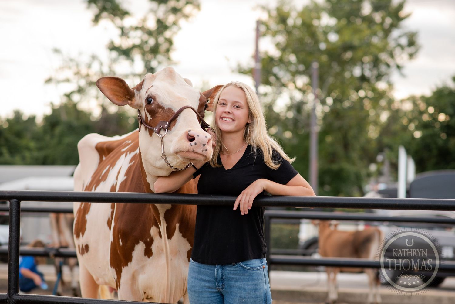 A brown and white dairy cow and handler pose together by a metal fence during golden hour.