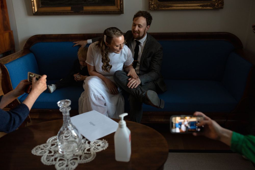 A wedding couple sits on a blue couch while others take photos during an intimate home ceremony.