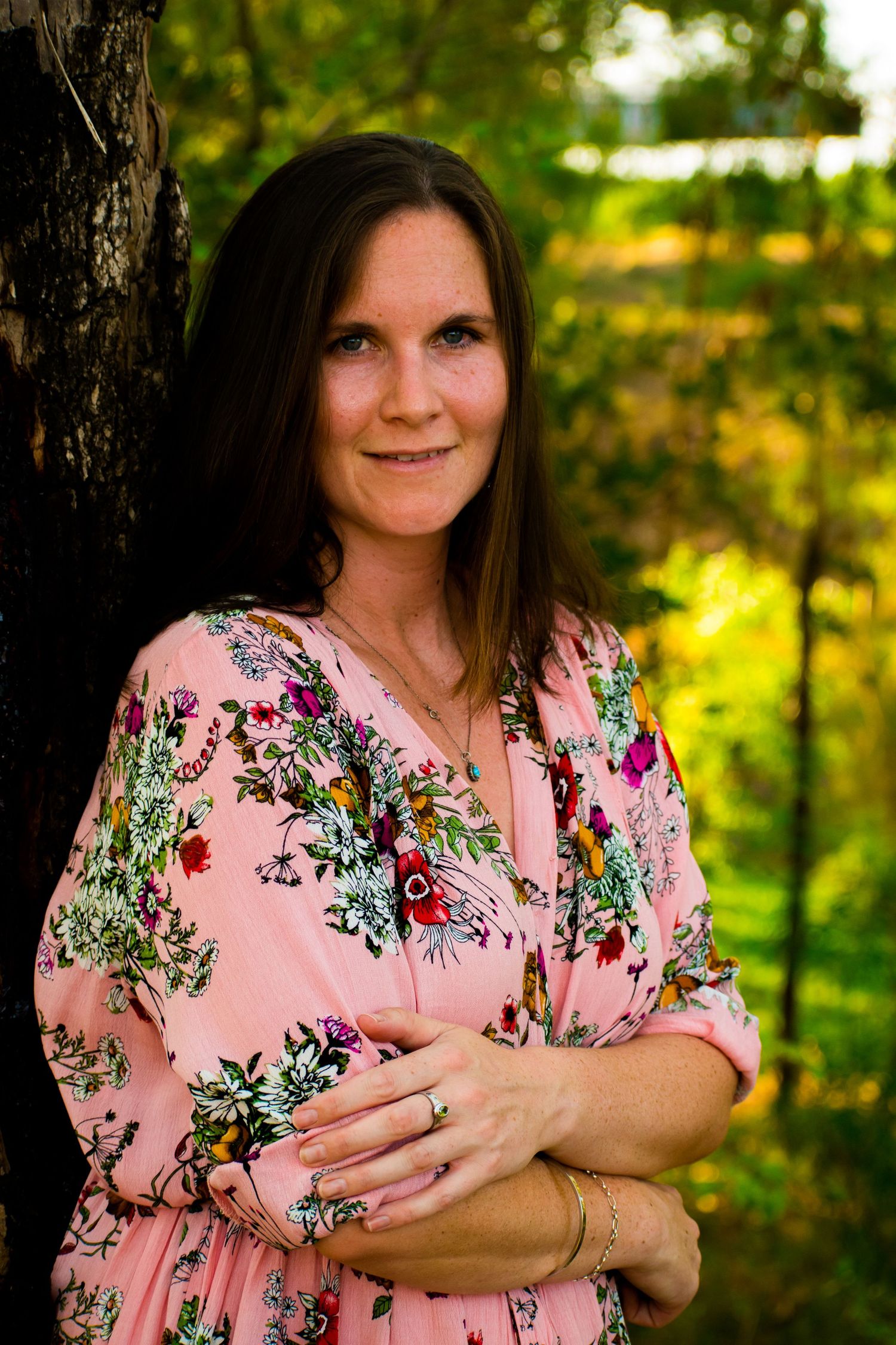 Woman in pink floral dress poses against sunlit forest background in outdoor portrait series.