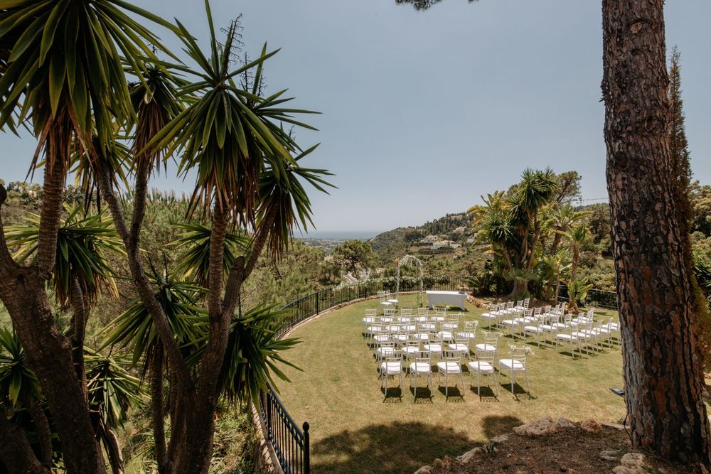 Outdoor wedding ceremony setup with white chairs on a grassy hillside surrounded by tropical palm trees.