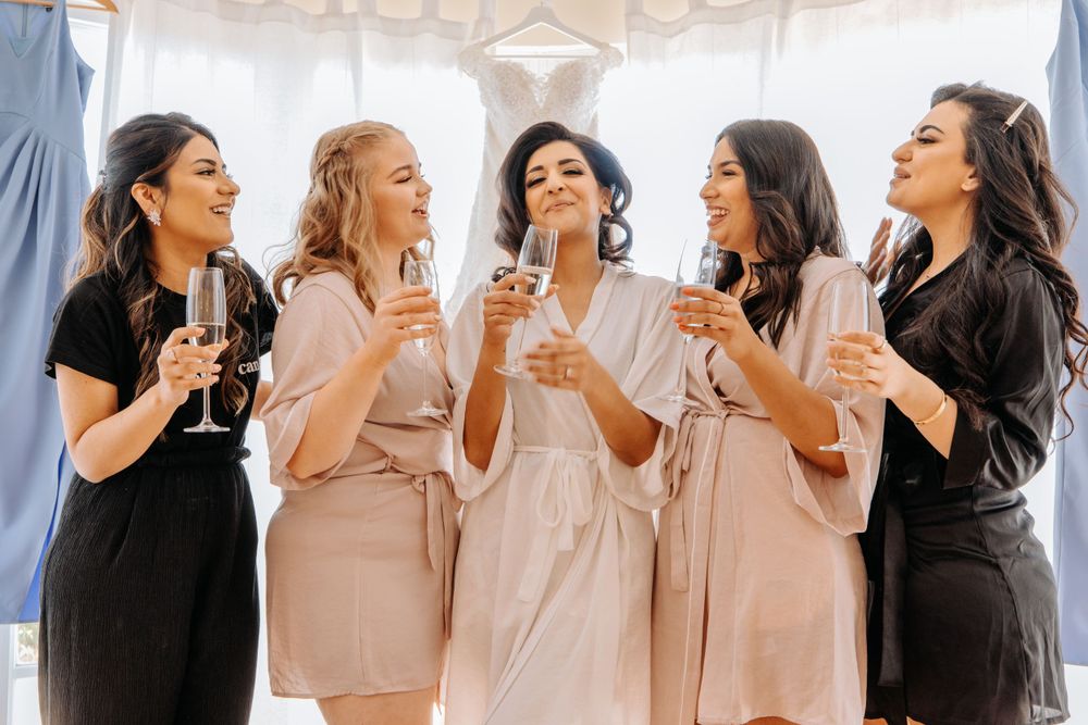 Women in matching robes laugh and toast with champagne glasses during wedding preparations.