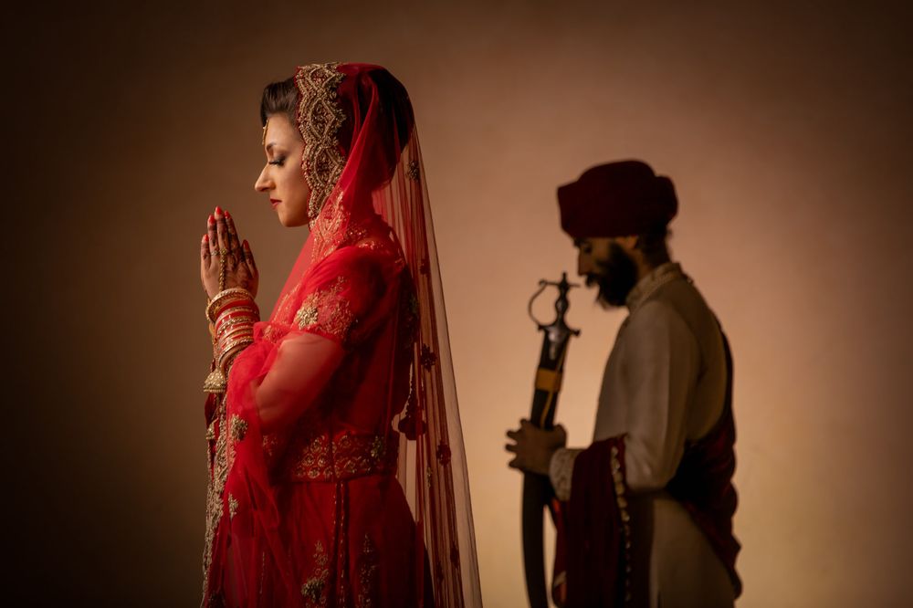 Artistic silhouette of bride in red veil praying while figure stands in background.