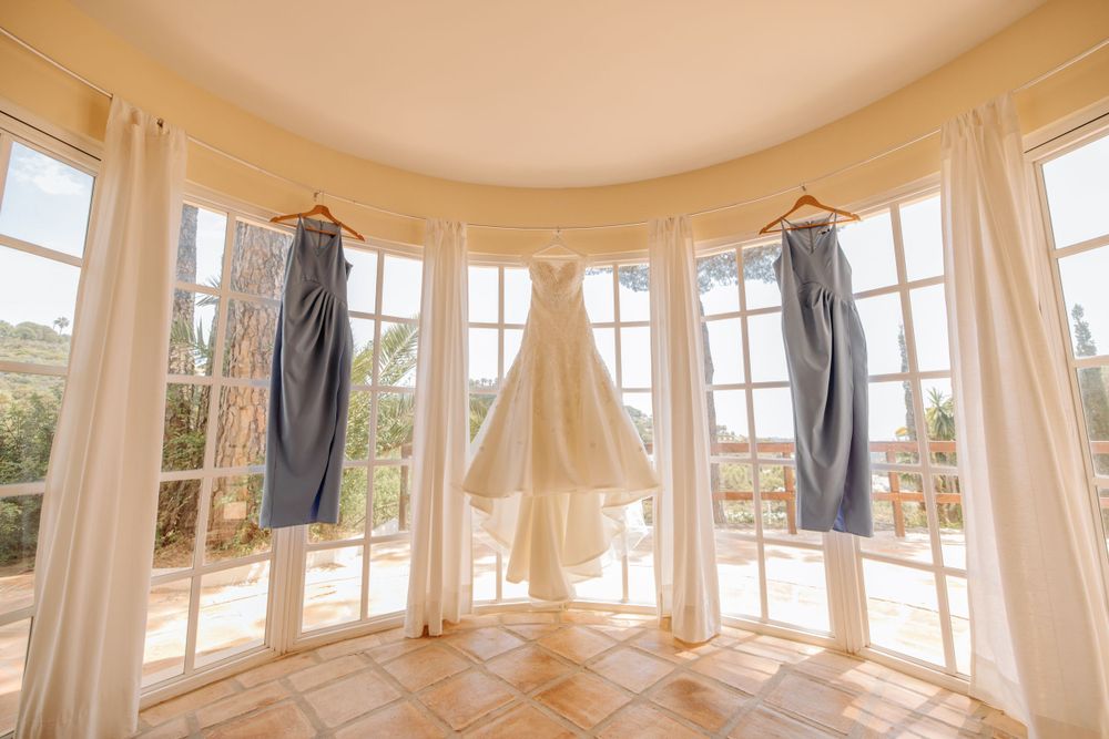 Wedding dress and blue suits hanging in bright bay window with natural lighting.