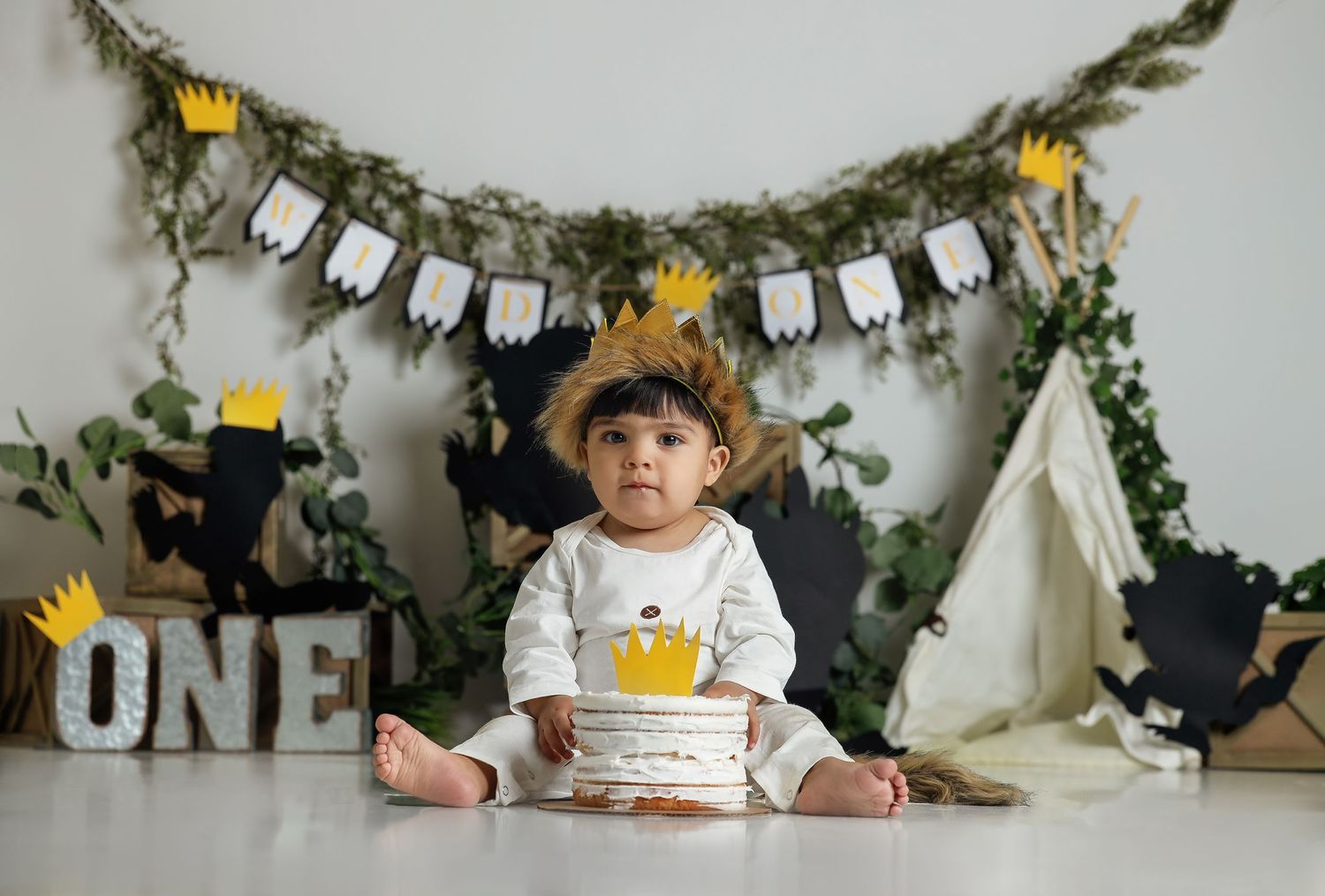First birthday setup with crown-wearing baby sitting against decorated wall with 'ONE' text.