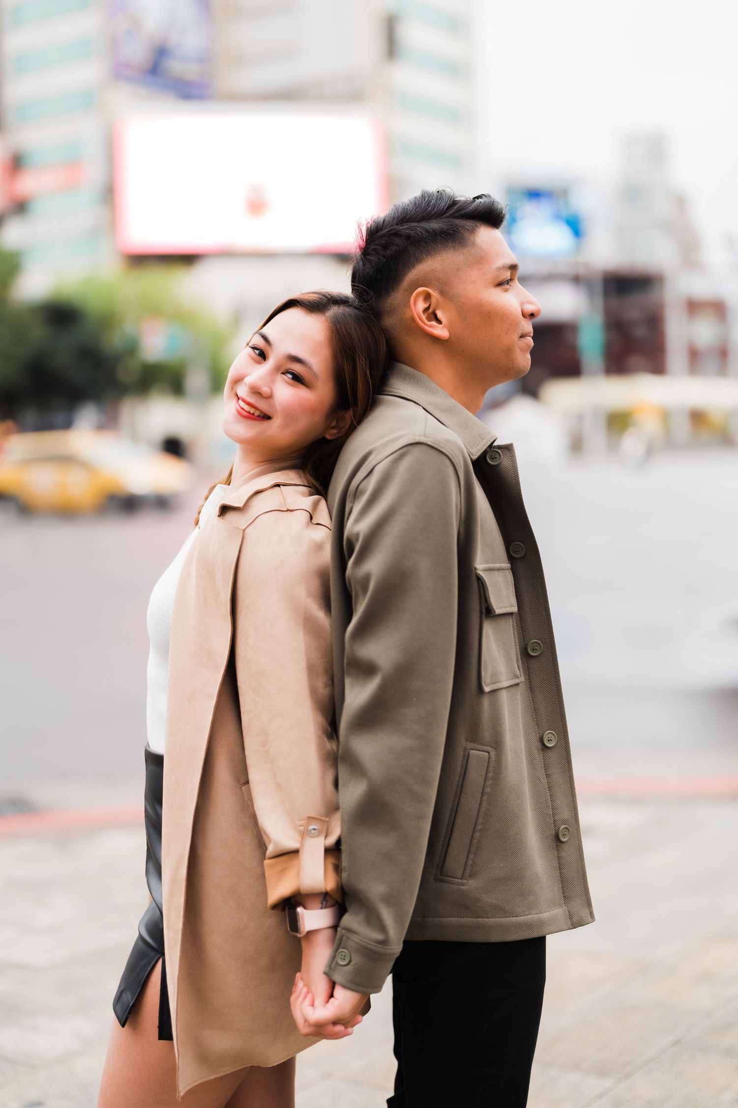 Stylishly dressed couple in matching neutral coats leaning back-to-back on a city street.