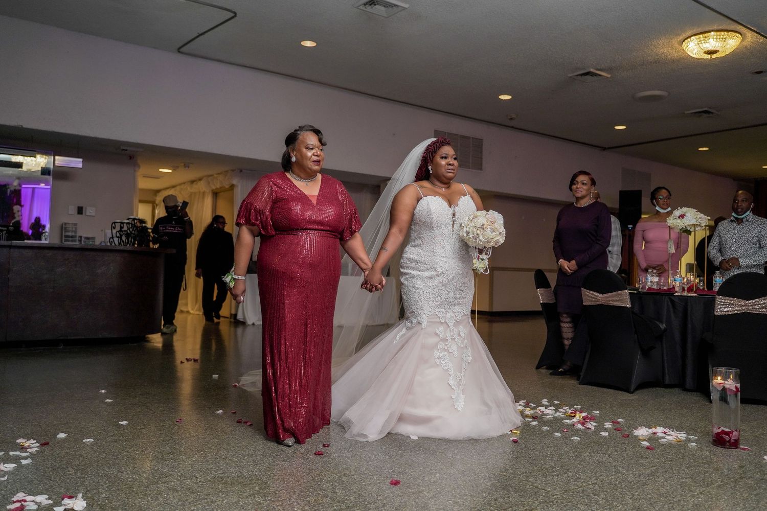 A wedding party stands in a reception hall with flower petals scattered on the floor.