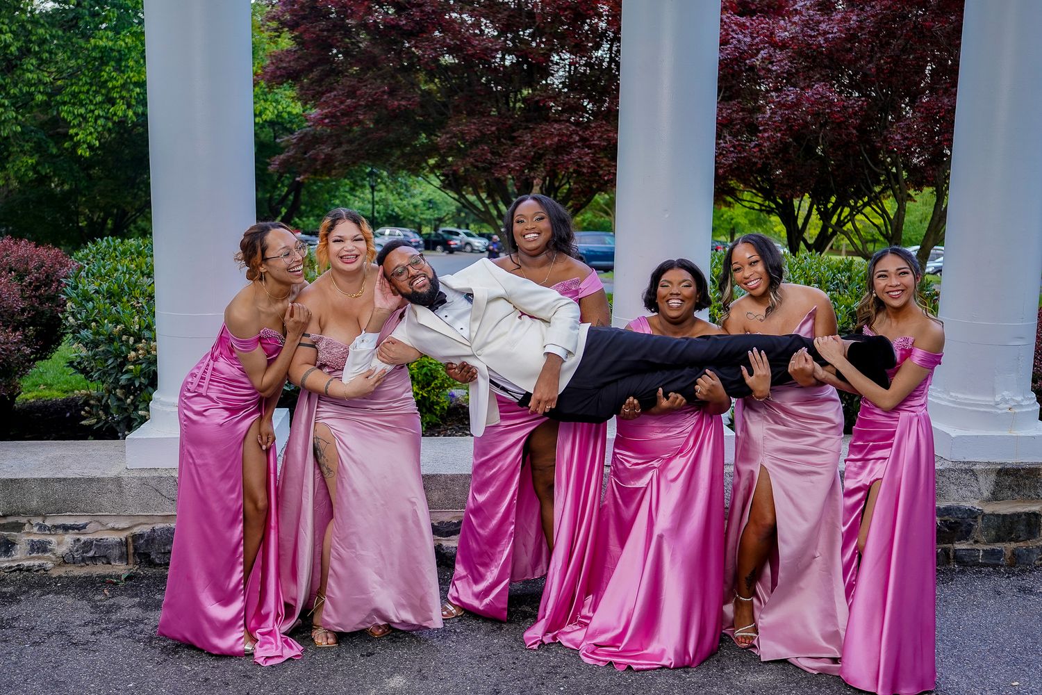 Wedding party in pink bridesmaid dresses poses under white columns outdoors.