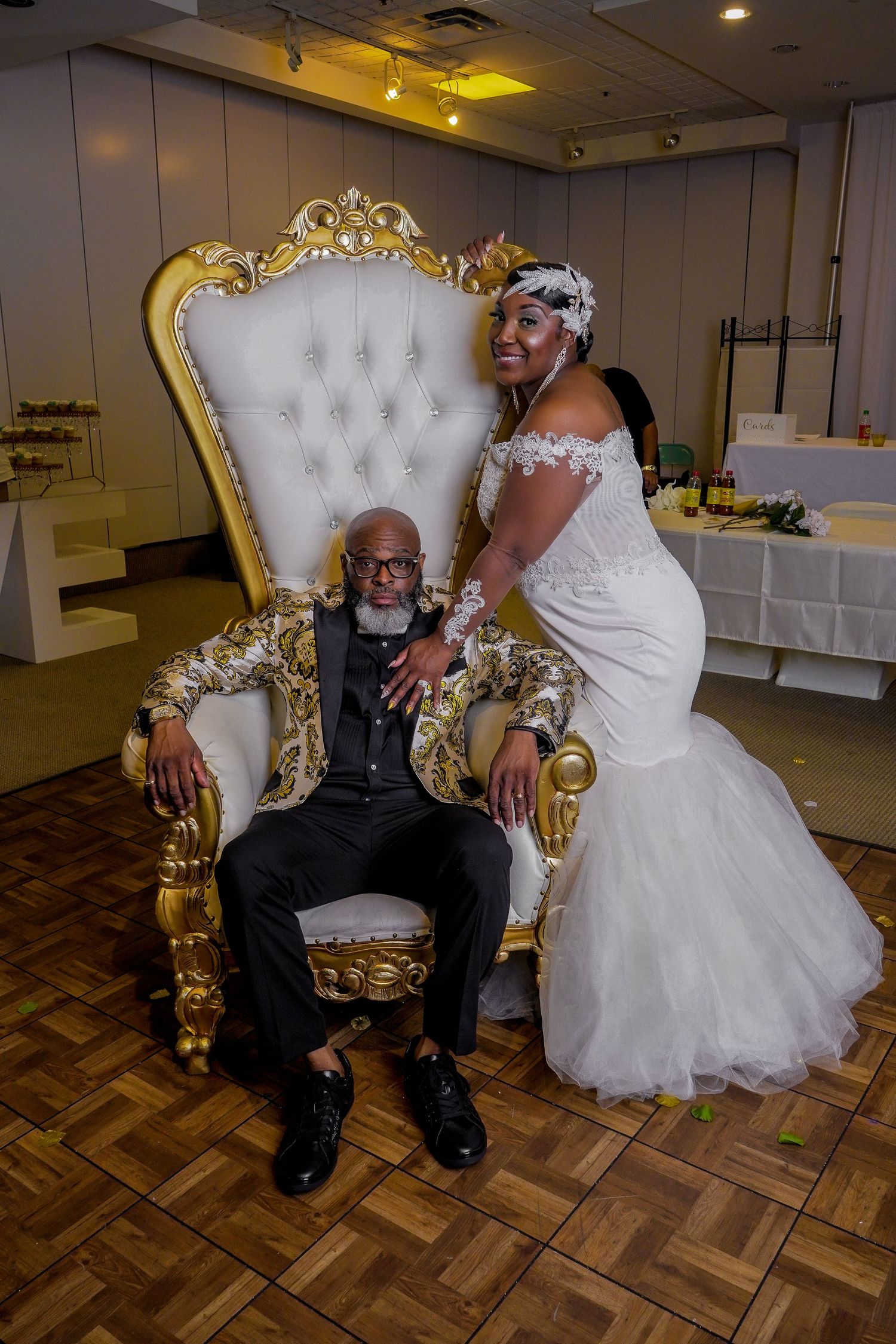 Wedding couple seated on ornate gold and white throne chair with parquet flooring.